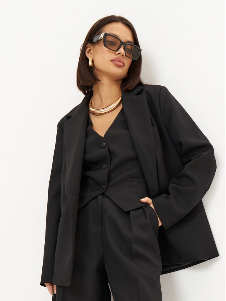 A woman confidently wearing large, black oversized square sunglasses, with a minimalist white blazer