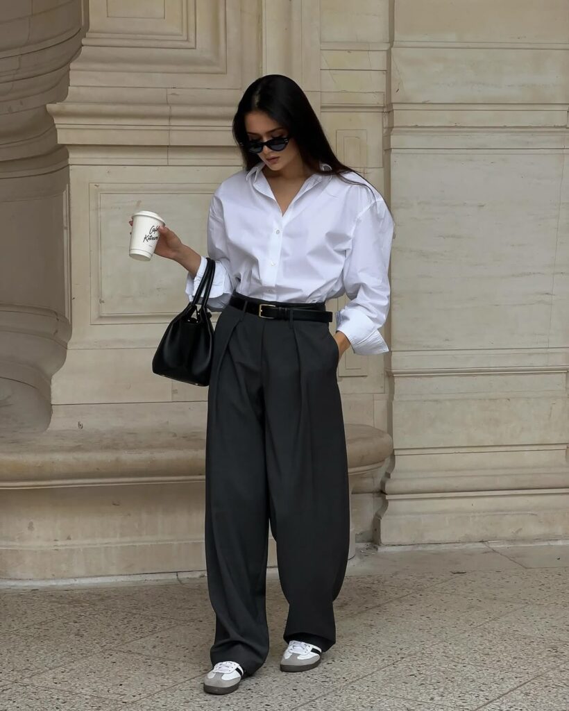 A flat lay of clean girl essentials including a white t-shirt, tailored black trousers, a simple gold necklace, a neutral handbag, and a pair of classic loafers